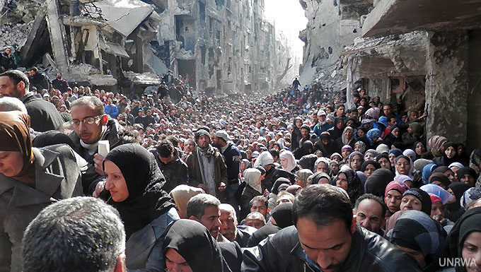 A gathering of people in a middle eastern town. Surrounding buildings appear to be crumbling and there is a large gathering in the middle of the street with innumerable people.